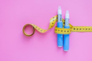 Blue peptide injection pens wrapped with yellow measuring tape on pink background, representing peptide therapy for weight loss and body measurement tracking