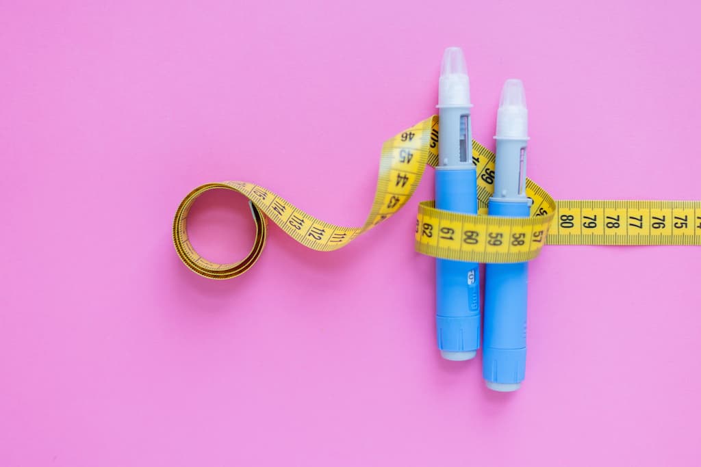 Blue peptide injection pens wrapped with yellow measuring tape on pink background, representing peptide therapy for weight loss and body measurement tracking